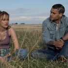 Zwei Personen sitzen auf einem Feld. Die Frau trägt ein lila Top, der Mann ein Jeanshemd. Beide schauen ernst.