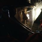 Ein Astronaut mit Helm und rotem Anzug untersucht etwas in einer dunklen Umgebung. Beleuchtung von oben.