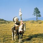Eine Person in traditioneller Kleidung reitet auf einem Pferd mit einer amerikanischen Flagge über eine weite Graslandschaft.
