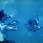 Unterwasseraufnahme: Ein Mann mit Sonnenbrille trinkt aus einer Flasche, während eine Frau in einem Bikini auf ihn zuschwimmt.