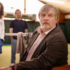 Ein Mann in Anzug sitzt in einer Tennishalle, zwei Personen stehen im Hintergrund. Ein Handtuch hängt am Netz.