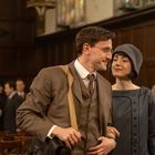 Ein Mann und eine Frau in eleganter, altmodischer Kleidung, sie lächelt ihn an; im Hintergrund Holzvertäfelung.