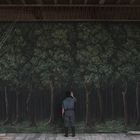 Eine Person steht vor einem großen gemalten Wald auf einer Theaterbühne, umgeben von Holz und Seilen.