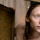 Eine junge Person mit nassen Haaren schaut durch einen Spalt in einer Holzwand, wirkt nachdenklich.