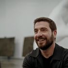 Ein Mann mit Bart lächelt in einem Raum mit einer großen Skulptur von Händen, die ein Herz formen, im Hintergrund.