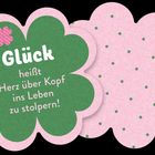 Text: "Glück heißt Herz über Kopf ins Leben zu stolpern!" Illustration in Kleeblattform, grün und rosa gestaltet.
