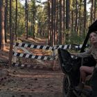 Schild: "ROAD CLOSED, VISITORS WELCOME". Frau sitzt in einem offenen Geländefahrzeug vor einer Barriere im Wald.