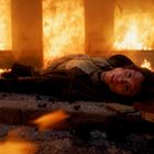 Ein Mensch liegt auf einem Boden, umgeben von Flammen und Trümmern, mit einem erschöpften Ausdruck im Gesicht.