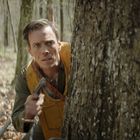 Ein Mann in einem gelben Schutzanzug späht wachsam hinter einem dicken Baumstamm in einem Wald hervor.