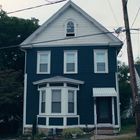 Zweistöckiges dunkelblaues Haus mit weißen Fensterrahmen und Ziegeldach, daneben ein Auto und Bäume im Hintergrund.