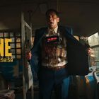 Text: "ONE," "09:59:59." Ein Mann in einem Bus zeigt unter seinem Anzug Sprengstoff mit einer digitalen Zeitanzeige.