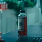 Auf einem Tisch steht eine Flasche mit der Aufschrift "Mother's Secret Kombucha". Im Hintergrund verschwommene Pflanzen.