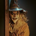 Ein gruseliger Vogelscheuchen-Clown schaut durch einen Türspalt, mit zerzaustem Hut und angsteinflößendem Gesicht.