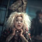 Eine Frau mit wildem, blondem Haar, aufgeregtem Gesichtsausdruck, trägt bunte, gemusterte Kleidung. Hintergrund unscharf.