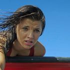 Ich kann keine sichtbaren Texte oder Hinweise erkennen. Eine Frau mit nassen Haaren blickt ernst unter blauem Himmel.