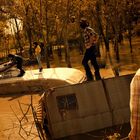 Vier Personen stehen auf einem versunkenen Wohnmobil in einem überfluteten Wald. Matschtöne dominieren die Szene.