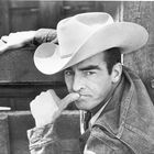 Ein Mann mit Cowboyhut und Jeansjacke lehnt nachdenklich an einem Holzgeländer, sein Finger berührt die Lippen.