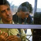 Zwei Männer in einer Telefonzelle. Einer schaut nachdenklich, der andere telefoniert intensiv. Draußen Landschaft.