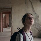 Keine Texte erkennbar. Eine Person mit Rucksack blickt in einer verlassenen, rissigen Betonumgebung nach oben.