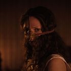 Eine Frau mit lockigem Haar trägt ein bronzenes, maskenartiges Gerät auf ihrem Gesicht. Der Hintergrund ist dunkel.