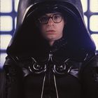 Ein Mensch mit Brille trägt einen großen, schwarzen Helm und eine Rüstung. Der Hintergrund hat helle, quadratische Muster.