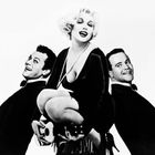 Ein humorvolles Schwarz-Weiß-Foto zeigt eine Frau zwischen zwei Männern, alle lächeln elegant und chic gekleidet.