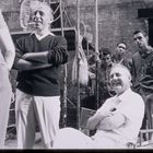 Mehrere Männer lächeln und sprechen auf einem Set, zwei stehen im Vordergrund, einer sitzt lachend. Rustikale Umgebung.