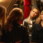 Zwei Frauen in glitzernder Kleidung schauen in einen Spiegel. Eine trägt silber, die andere schwarz.