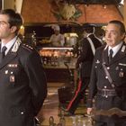 Zwei Männer in Uniform stehen in einem Restaurant, im Hintergrund ein Koch und Flaschen im Regal.