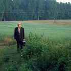 Ein Mann in Anzug mit Aktentasche steht auf einem grünen Feld vor einem Wald.