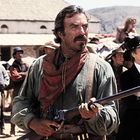 Ich sehe keinen Text. Ein Mann in Westernkleidung hält ein Gewehr, umgeben von Leuten in einer wüstenartigen Szenerie.