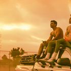 Zwei junge Männer sitzen ohne Hemd auf einem Auto, der Himmel ist orange gefärbt.