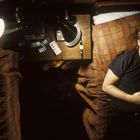 Ein Mann liegt nachdenklich auf einem Bett, im Zimmer stehen Lampen, ein Tisch mit Kassetten und eine Fotografie.