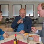 Ein Mann in einem Anzug isst Eintopf. Zwei weitere Personen sitzen an einem Tisch in einer Kantine.