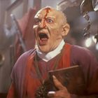 Eine Figur in rotem Gewand mit schreckenserfülltem Gesichtsausdruck, Blut läuft über die Stirn. Im Hintergrund eine Statue.