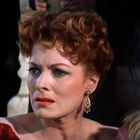 Eine Frau mit roten Haaren und besorgtem Ausdruck trägt ein rotes Kleid und goldene Ohrringe.