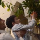 Ein Mann hebt ein Kind, das eine Orange von einem Baum pflückt, beide lächeln. Im Hintergrund sind Gebäude zu sehen.