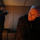 Ein Mann mit besorgtem Gesichtsausdruck befindet sich im Vordergrund. Im Hintergrund zielt eine Person mit einem Gegenstand auf ihn.