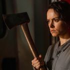 Keine erkennbaren Texte. Eine Frau mit ernstem Gesichtsausdruck hält eine Axt, im Hintergrund ein roter Lichtschein.