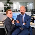 Zwei Männer in Anzügen. Einer schaut nervös. Im Hintergrund Büro mit Computer und Papieren. "Capitol" ist lesbar.