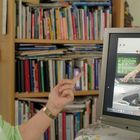 Eine Frau zeigt auf einen Monitor mit einem Video. Im Hintergrund ist ein Bücherregal zu sehen.