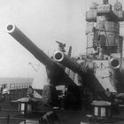 Zwei große Kanonenrohre eines Kriegsschiffs, mit einem Turm im Hintergrund. Keine sichtbaren Texte.