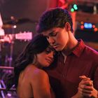 Ein Paar tanzt eng umschlungen, während im Hintergrund eine Person Gitarre spielt. Die Atmosphäre ist warm und intim.