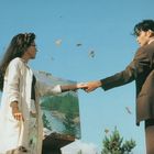 Ein Mann und eine Frau stehen vor einem Himmel, halten Hände, umgeben von Schmetterlingen; eine Staffelei zeigt eine Landschaft.