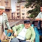 Keine erkennbaren Texte. Vier Personen unterhalten sich draußen vor einem Gebäude.