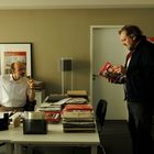 Zwei Männer in einem Büro, einer sitzt am Schreibtisch, der andere steht und hält eine Zeitschrift. Viel Papierkram.