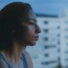 Keine Texte vorhanden. Ein Mensch blickt nachdenklich aus einem Fenster mit unscharfem Stadtgebäude im Hintergrund.