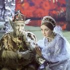 Ein älterer Mann mit Krone und Pelzmantel sitzt neben einer Frau in blauem Kleid mit Schmuck. Hintergrund in Rottönen.