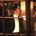 Ich kann den Text nicht erkennen. Eine Person in formeller Kleidung steht vor einem beleuchteten Fenster und schaut direkt.