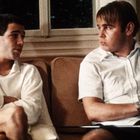 Zwei Männer sitzen auf einem Sofa, einer hält ein Gewehr. Beide tragen weiße Kleidung. Im Hintergrund ist ein Fenster.